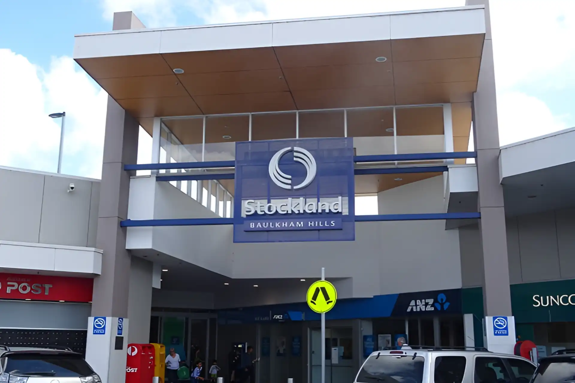 Entrance of the Stockland Baulkham Hills shopping centre with visible signage, a pedestrian crossing sign, and people walking around