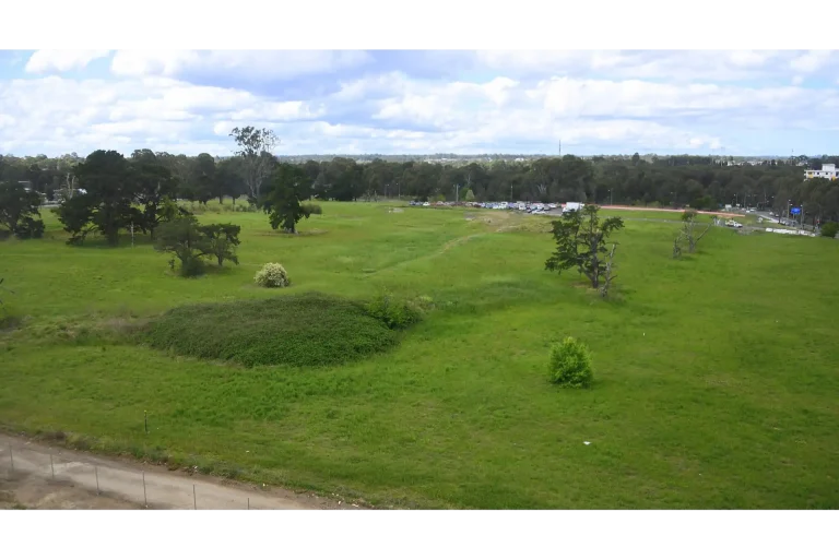 Expansive green field with scattered trees and a distant parking lot under a partly cloudy sky, viewed from an elevated perspective in Rouse Hill