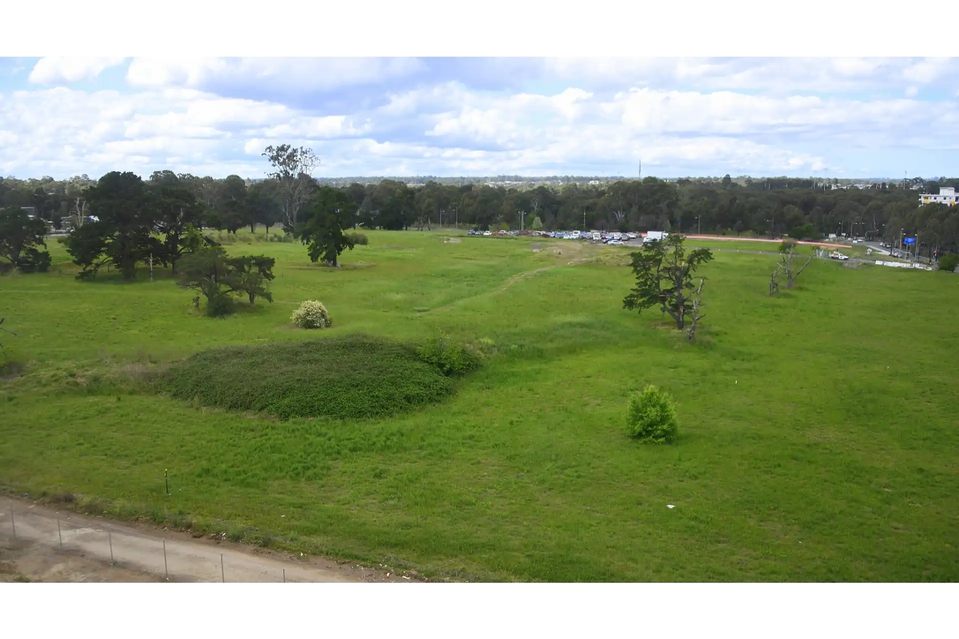 Expansive green field with scattered trees and a distant parking lot under a partly cloudy sky, viewed from an elevated perspective in Rouse Hill
