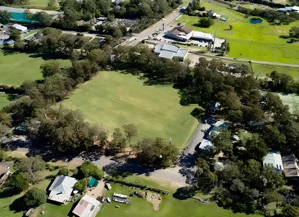 Aerial view of a spacious green field surrounded by trees with several buildings and a parking area nearby, set in a rural landscape.