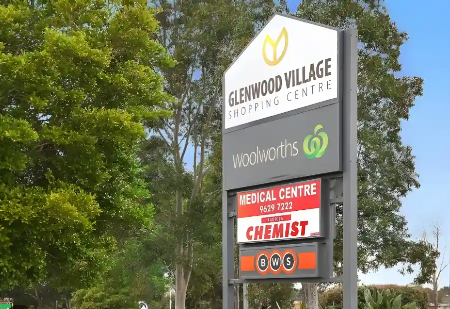 Signage post for Glenwood Village Shopping Centre with logos of Woolworths, a medical centre, a chemist, and BWS against a backdrop of green trees